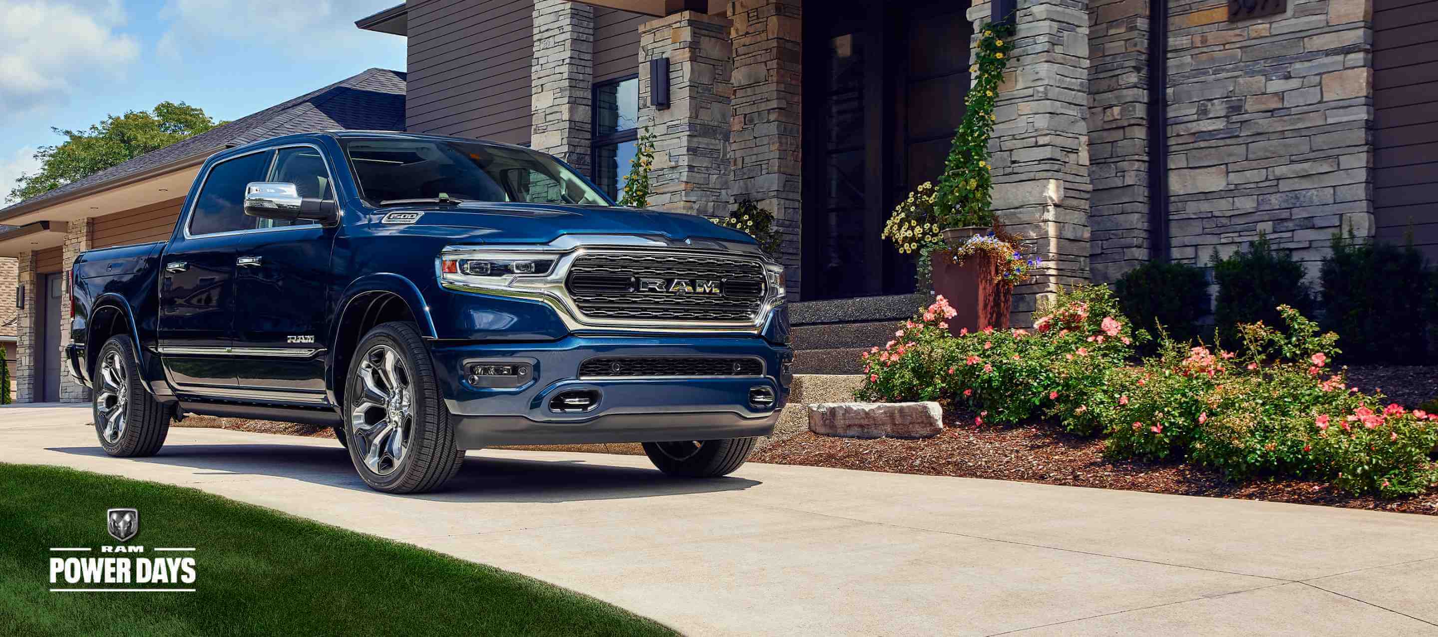 A front angled passenger-side profile of a blue 2024 Ram 1500 Limited Crew Cab parked in the driveway of a large home. Power Days Sales Event.