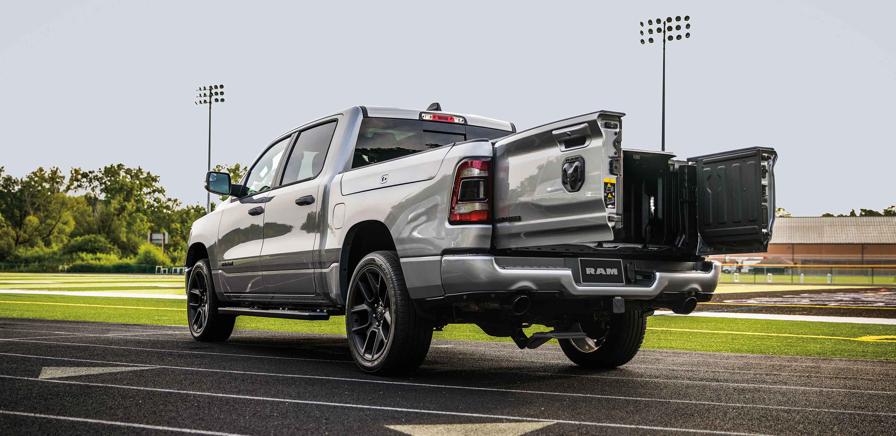 Display A silver 2024 Ram 1500 Laramie Crew Cab with the swing-open tailgate doors open, parked on a track beside a football field.