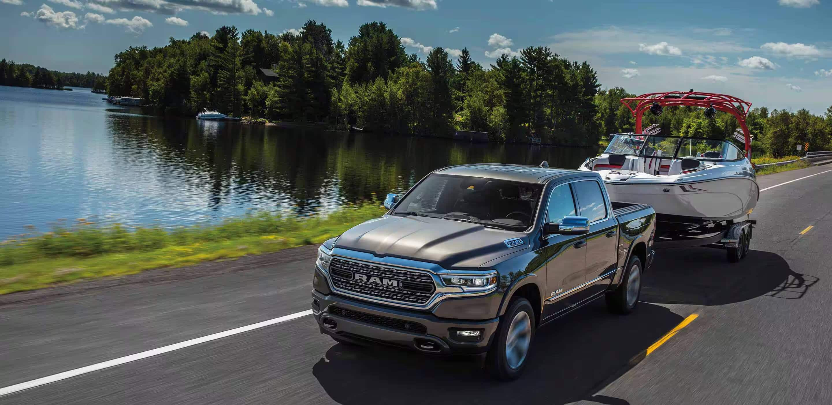 Display A silver 2024 Ram 1500 Limited Crew Cab towing a speedboat as it is driven down a highway beside a lake.