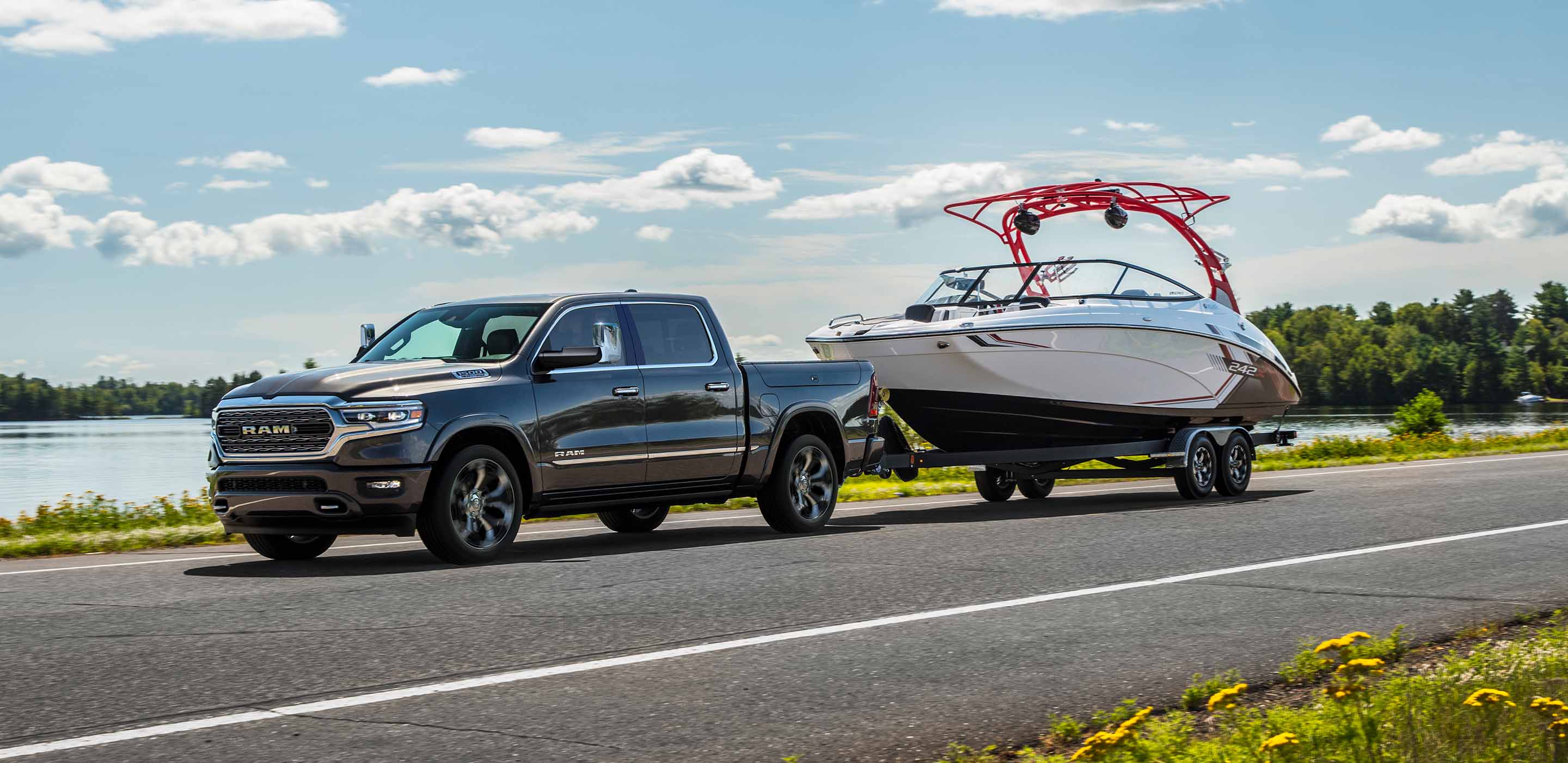 A silver 2024 Ram 1500 Limited Crew Cab towing a speedboat as it is driven down a highway beside a lake.