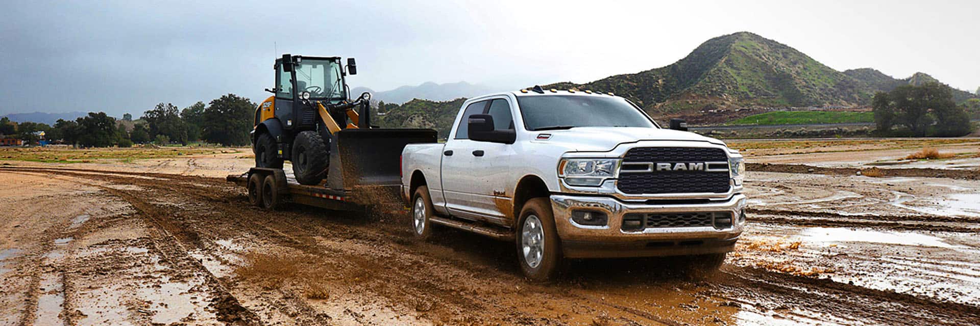 A white 2024 Ram 2500 Big Horn Crew Cab towing a front end loader on a flatbed trailer through a muddy construction site.