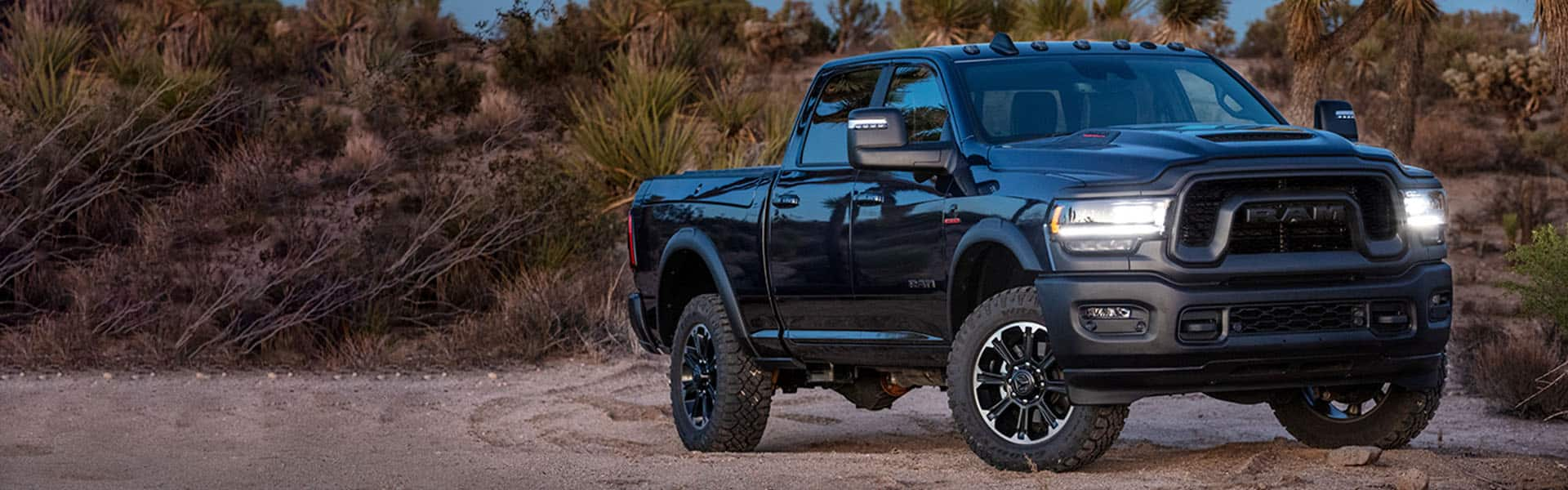 A 2024 Ram 2500 Rebel with its headlamps on, being driven off-road on a sandy trail.