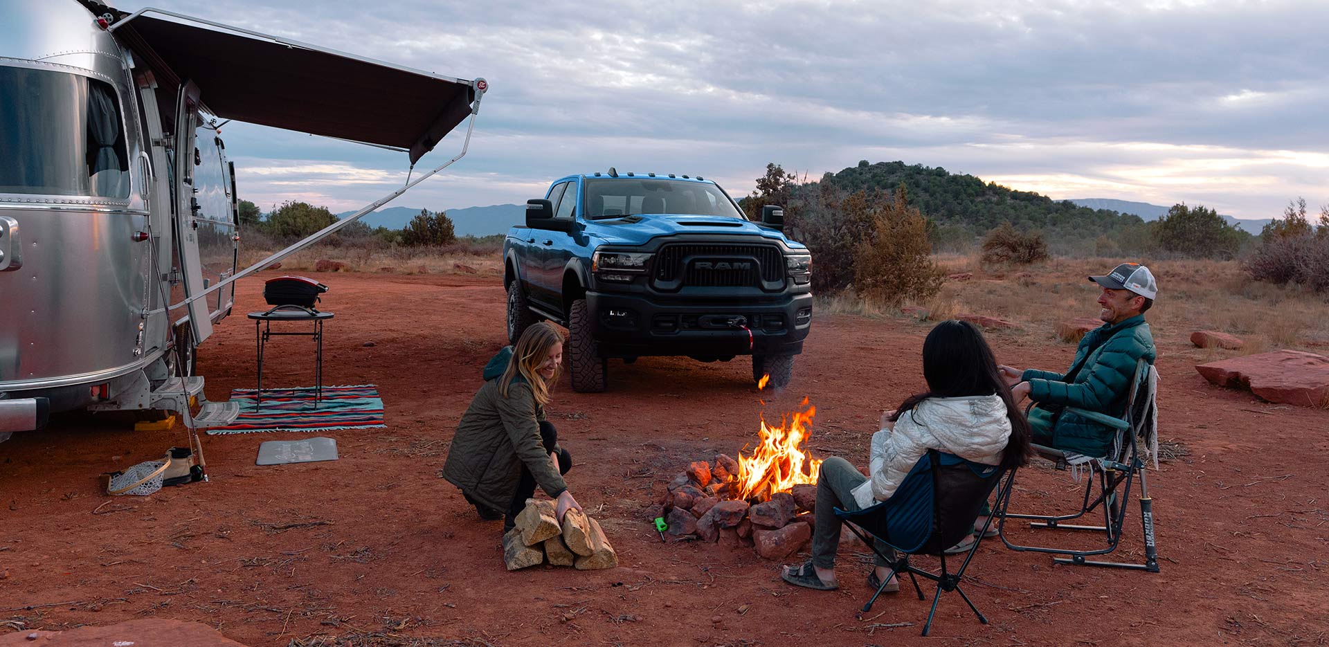 Display A blue 2024 Ram 2500 Power Wagon Crew Cab parked at a campsite in the desert, beside a camper trailer and three people sitting around a campfire.