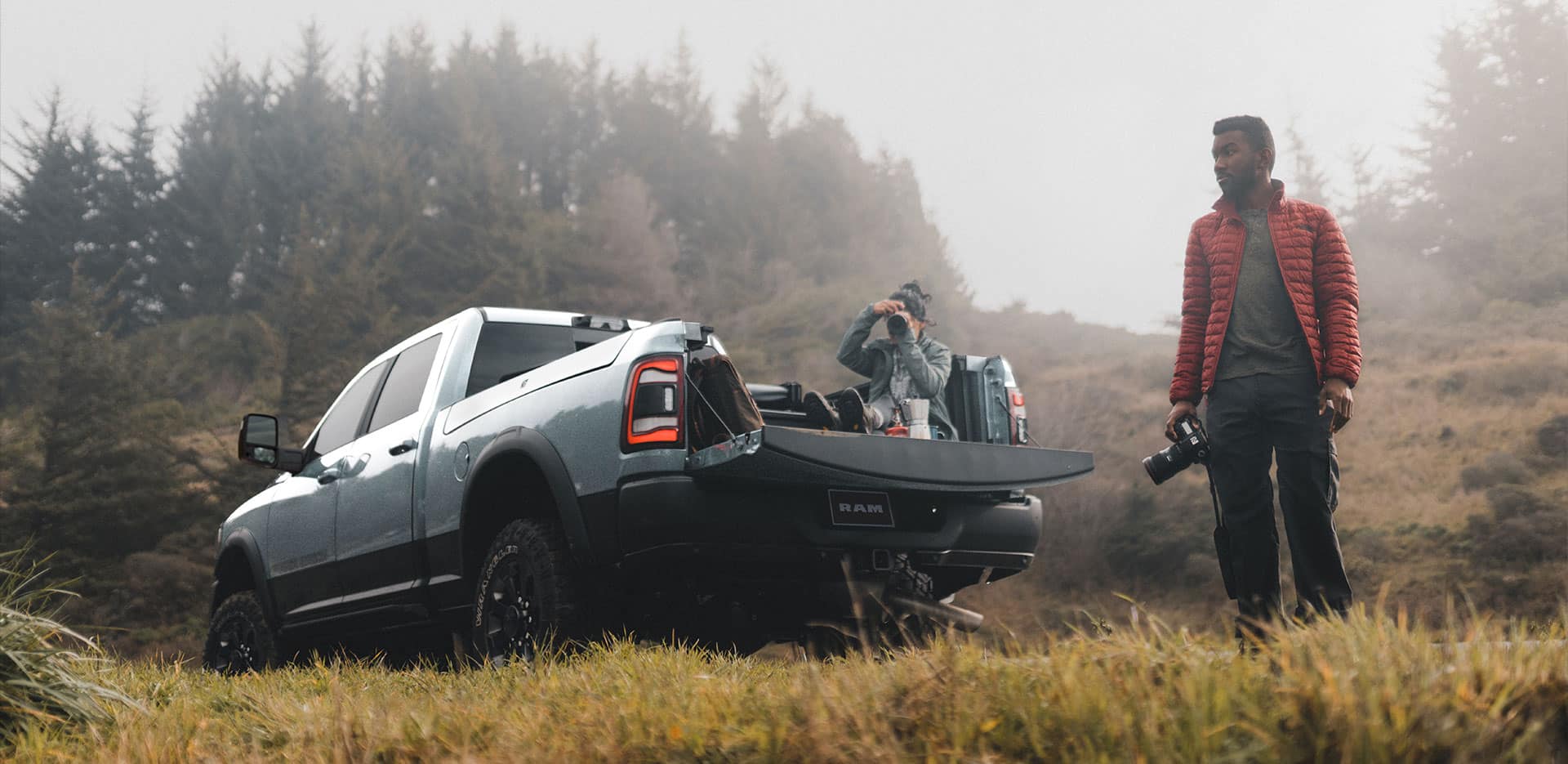 Display A 2024 Ram 2500 Power Wagon Crew Cab parked on a downward angle with its tailgate open, a person sitting in the pickup bed taking a photograph and another person standing beside the truck.