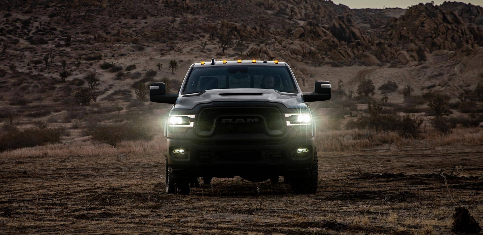 Display A head-on view of a 2024 Ram 2500 Rebel with its headlamps on, being driven off-road in the desert at dusk.