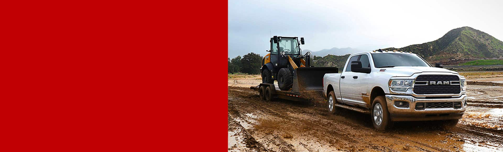 A white 2024 Ram 2500 Big Horn Crew Cab towing a front end loader on a flatbed trailer through a muddy construction site.