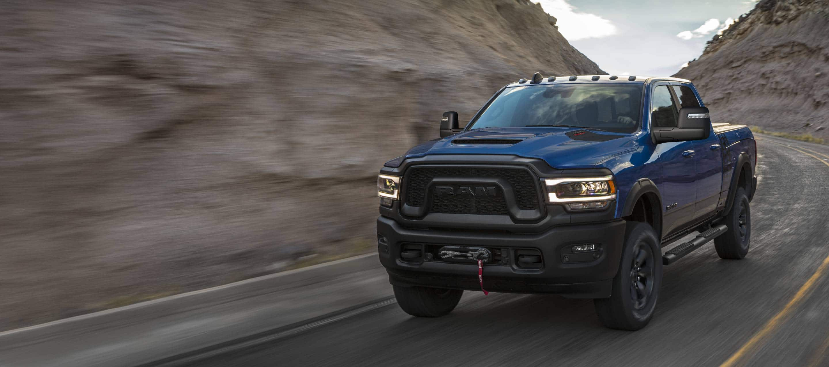 A blue 2024 Ram 2500 Power Wagon 4x4 Crew Cab being driven on a winding highway with hills on both sides. The background is blurred to indicate the vehicle is in motion.