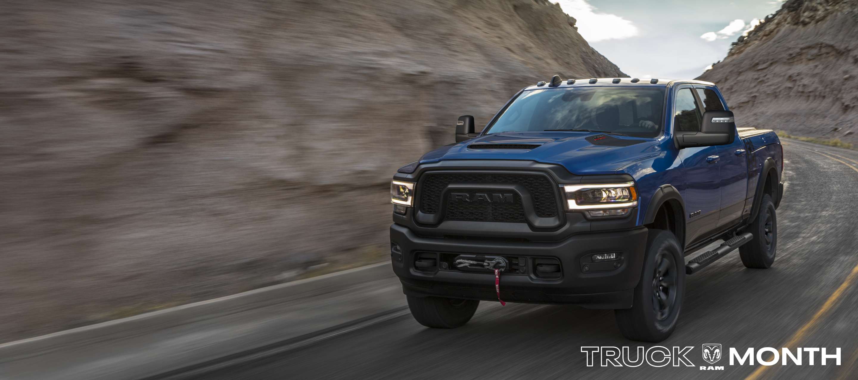 A blue 2024 Ram 2500 Power Wagon 4x4 Crew Cab being driven on a highway in the mountains. The background is blurred to indicate the truck is in motion. Truck Month Sales Event.