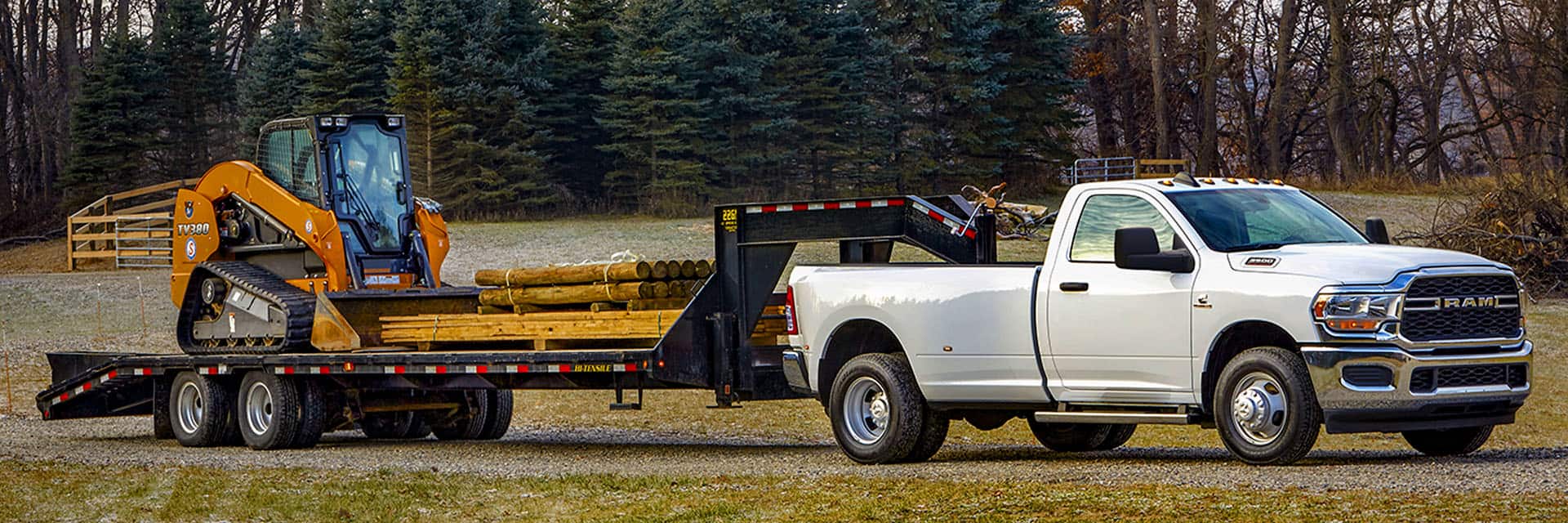 A white 2024 Ram 3500 Tradesman 4x4 Regular Cab towing a fifth wheel flatbed trailer loaded with an excavator and lumber.