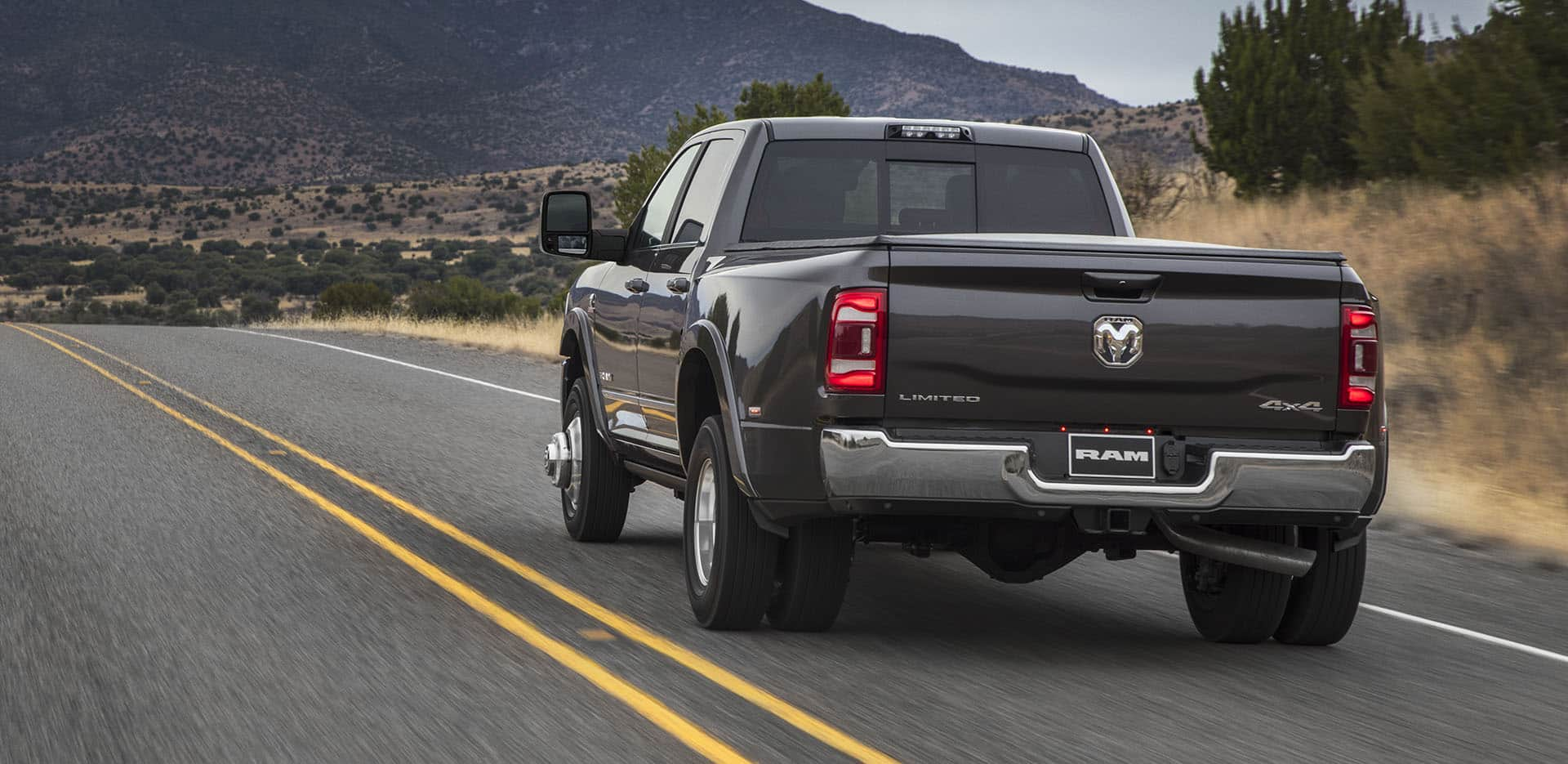 Display A rear angle of a gray 2024 Ram 3500 Limited Crew Cab being driven on a highway toward a group of mountains.