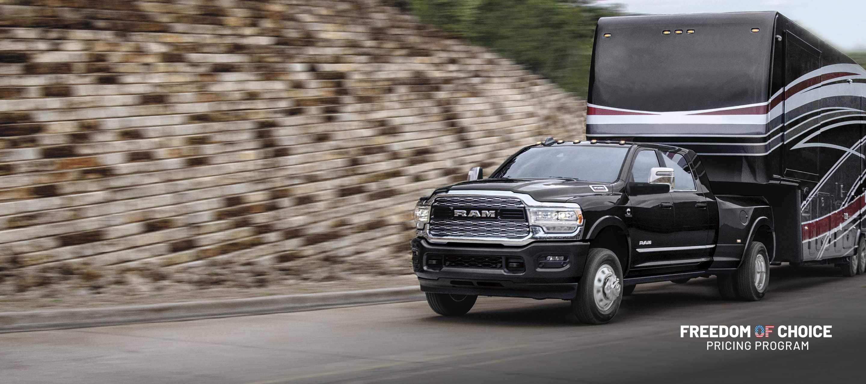 A black 2024 Ram 3500 Limited Mega Cab traveling down a highway, towing a large fifth wheel travel trailer. The background is blurred.  Freedom Of Choice Pricing Update Sales Event.
