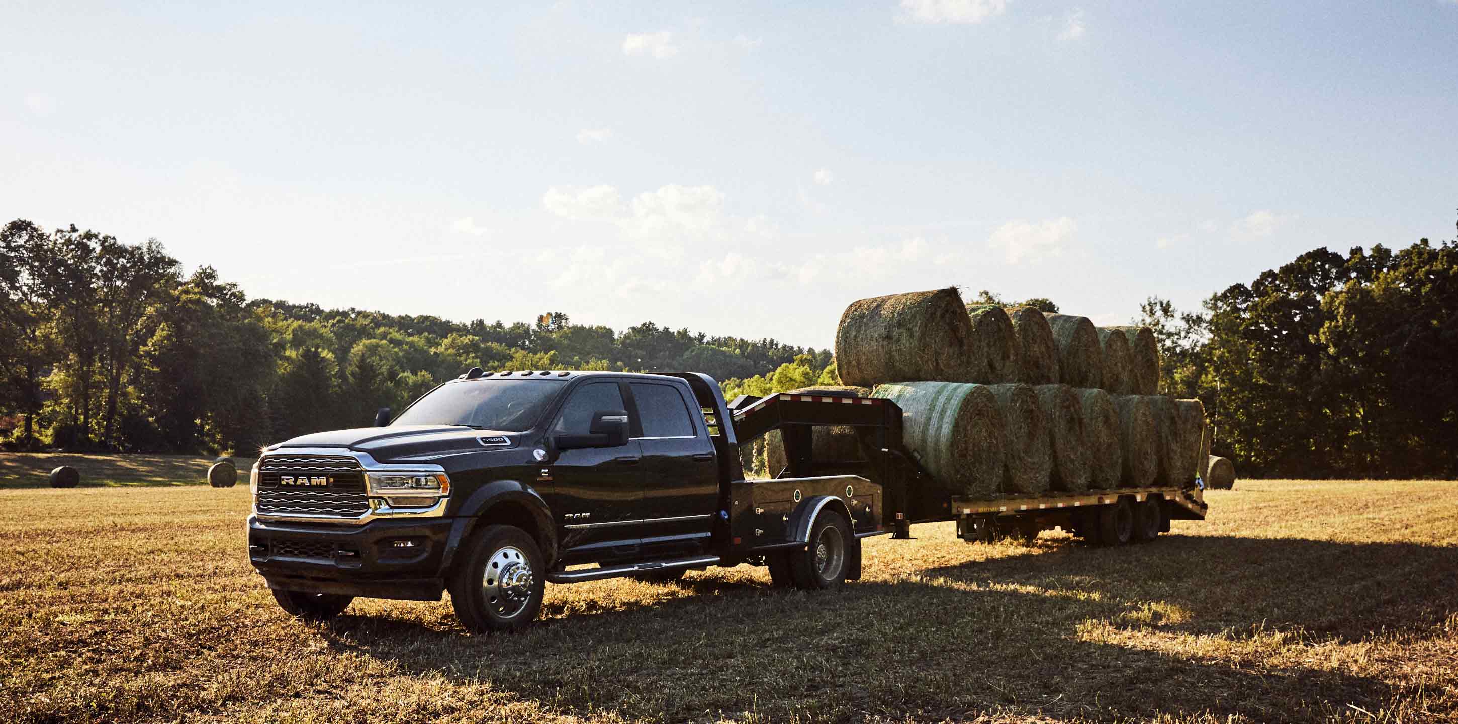 A black 2024 Ram 5500 Limited Chassis Cab Crew Cab with a gooseneck trailer loaded with several bales of hay, parked in a field.