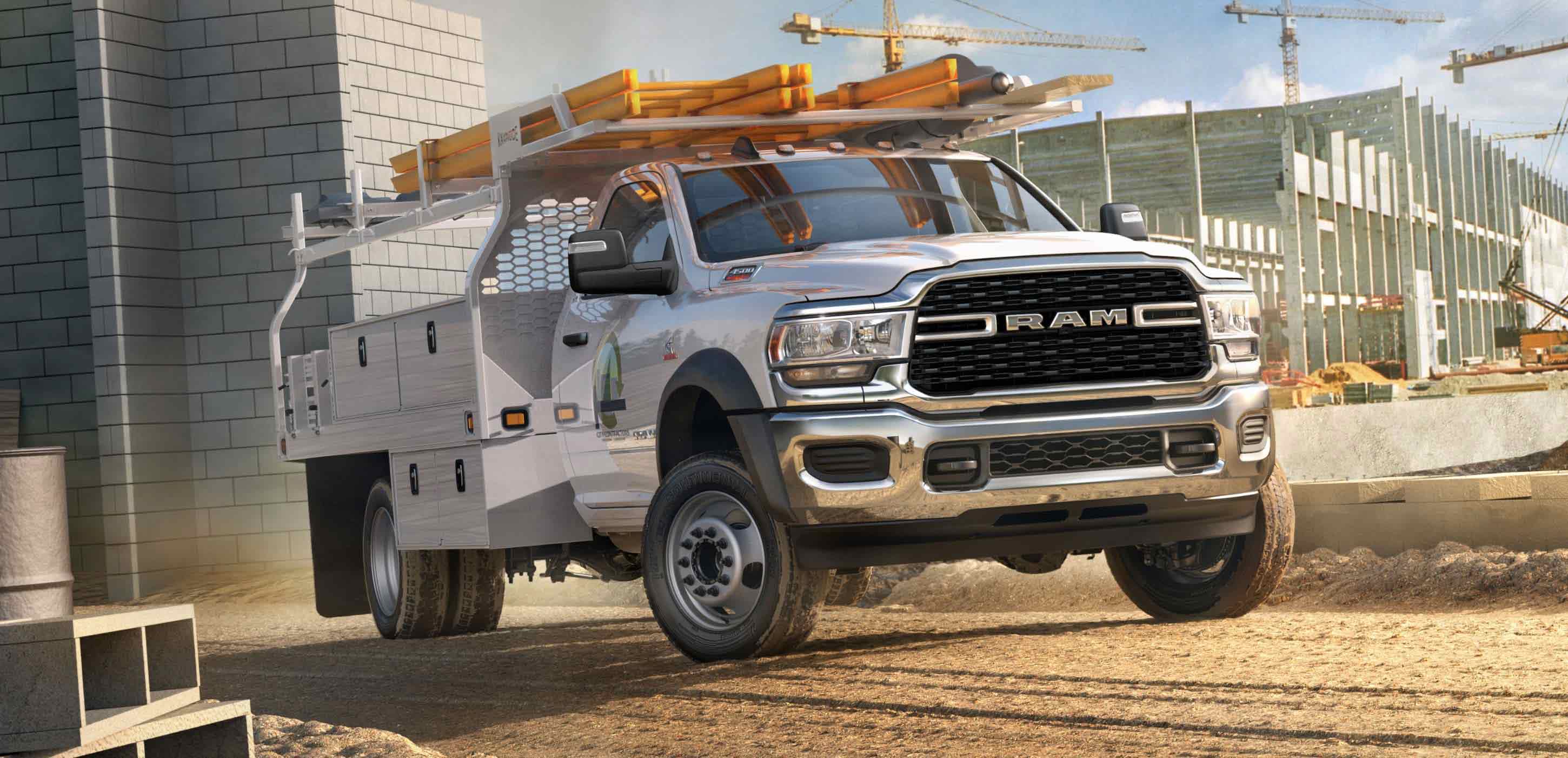 Display A white 2024 Ram 4500 SLT Chassis Cab Regular Cab with a utility upfit and ladder rack with two ladders attached, parked at a construction site.