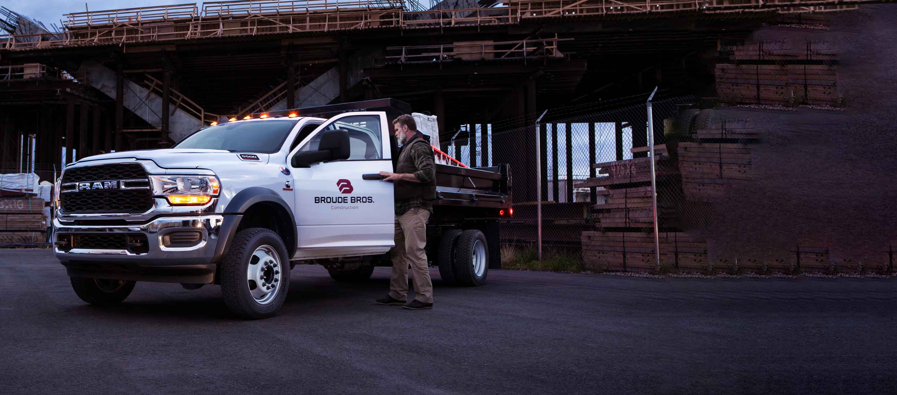 A man getting into the cabin of a white 2024 Ram 5500 Tradesman Chassis Cab Crew Cab with a stake bed upfit loaded with construction materials.