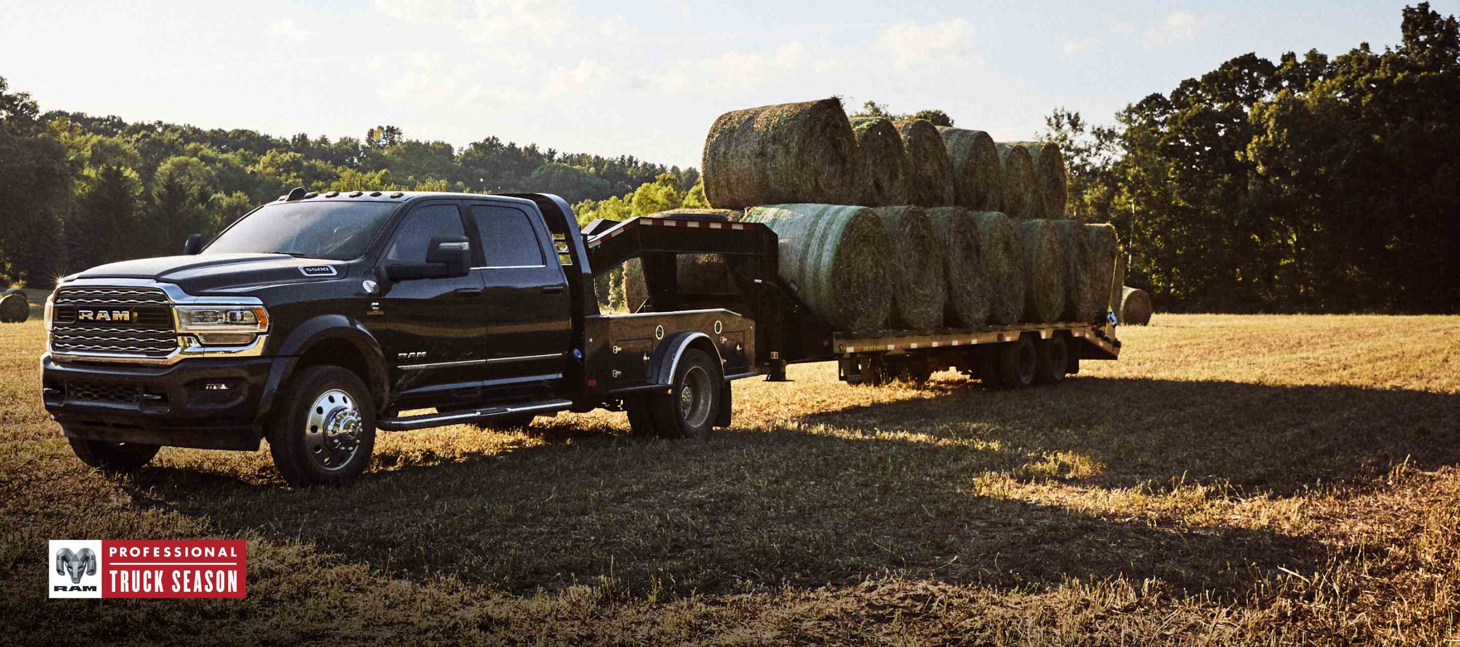 A black 2024 Ram 5500 Limited 4x4 Chassis Crew Cab with a gooseneck trailer loaded with several bales of hay, parked in an open field. Ram Professional Truck Season.