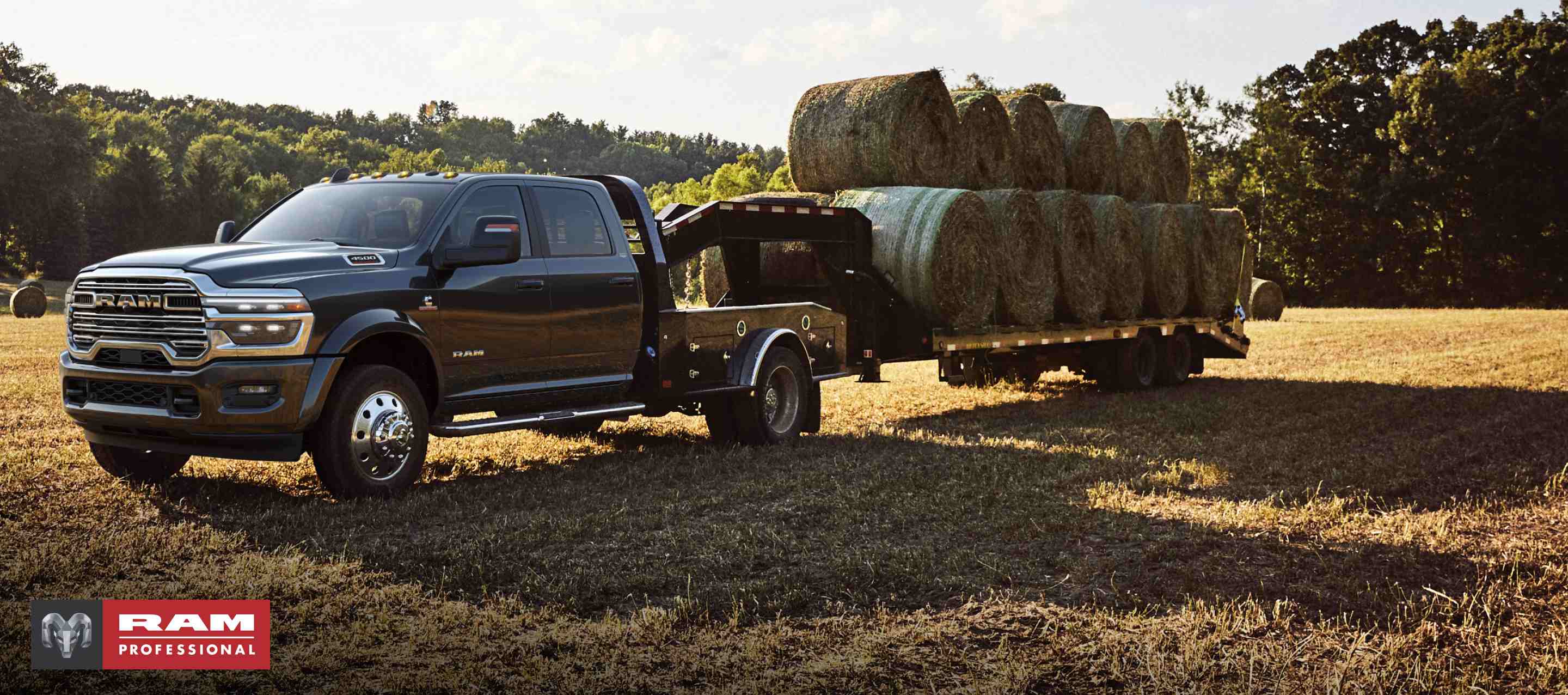 A black 2024 Ram 5500 Limited 4x4 Chassis Crew Cab with a gooseneck trailer loaded with several bales of hay, parked in an open field. Ram Professional.