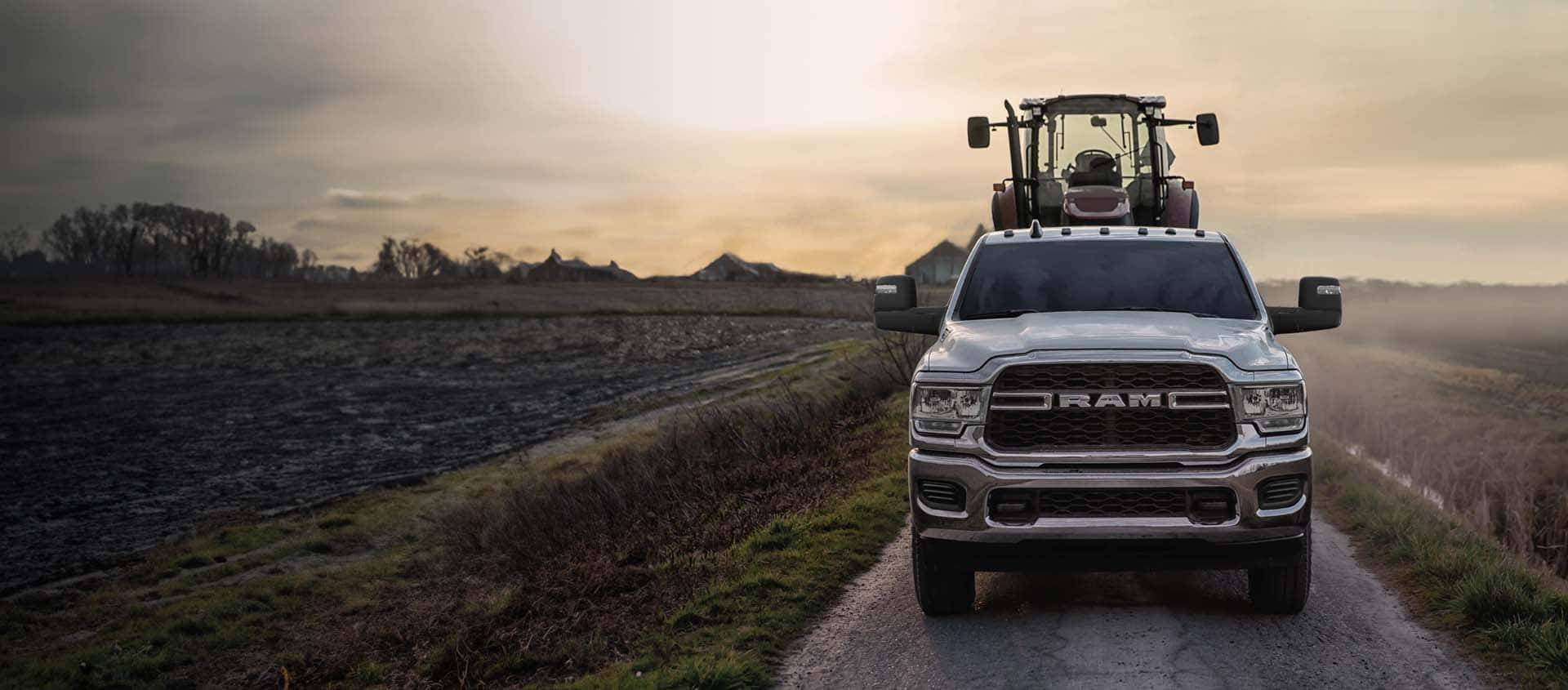 A 2024 Ram 2500 Tradesman Crew Cab  being driven down a single-lane country road, towing a construction vehicle.