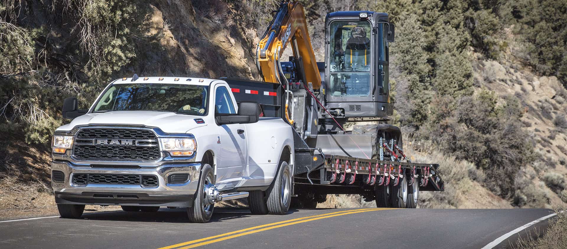 A white 2024 Ram 3500 Tradesman Regular Cab towing a fifth wheel flatbed trailer with an excavator on it.
