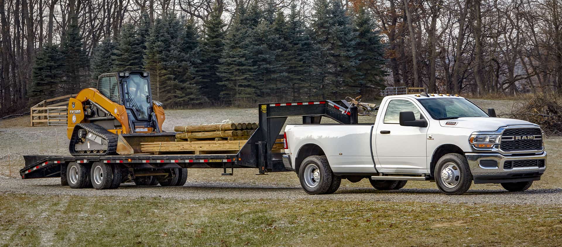 A white 2024 Ram 3500 Tradesman 4x4 Regular Cab towing a fifth wheel flatbed trailer loaded with an excavator and lumber. 