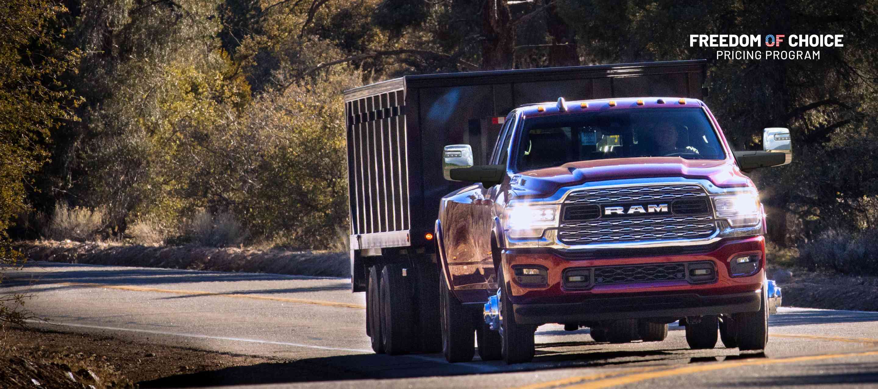 A red 2024 Ram 3500 4x4 Crew Cab traveling on a highway in the woods towing a dump body trailer. Freedom Of Choice Pricing Update Sales Event.