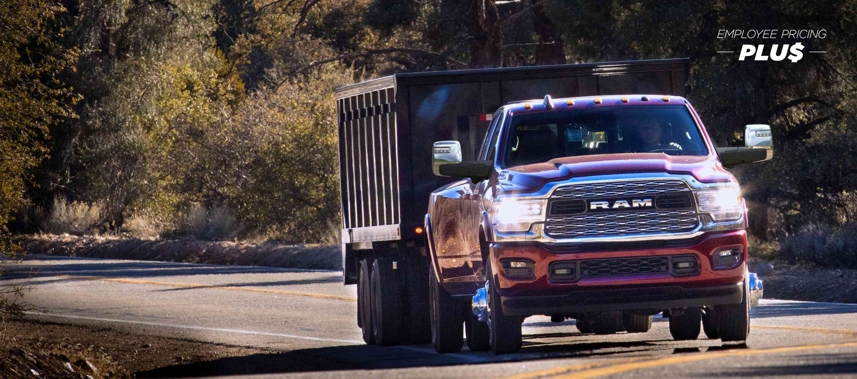 A red 2024 Ram 3500 4x4 Crew Cab traveling on a highway in the woods towing a dump body trailer. Employee Pricing Plus Sales Event.