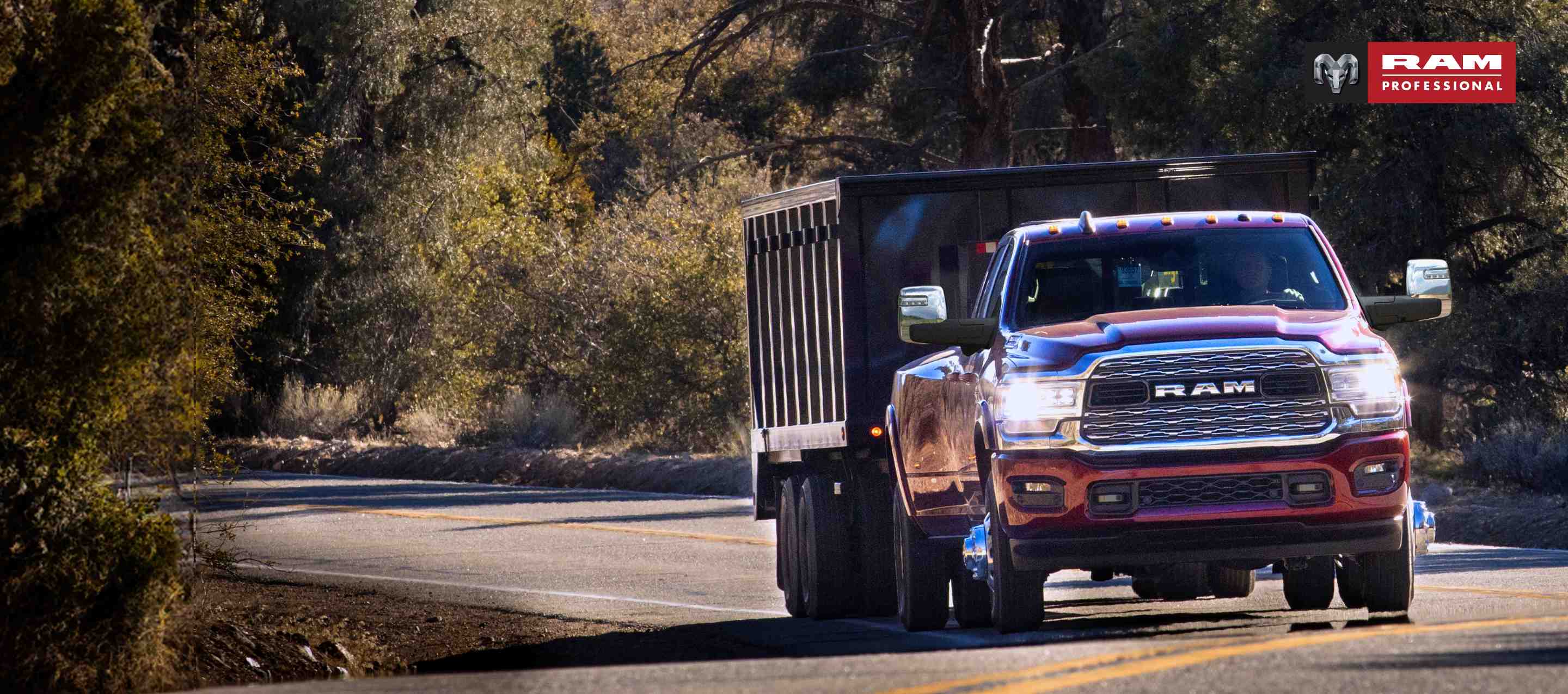 A red 2024 Ram 3500 4x4 Crew Cab traveling on a highway in the woods towing a dump body trailer. Ram Professional.