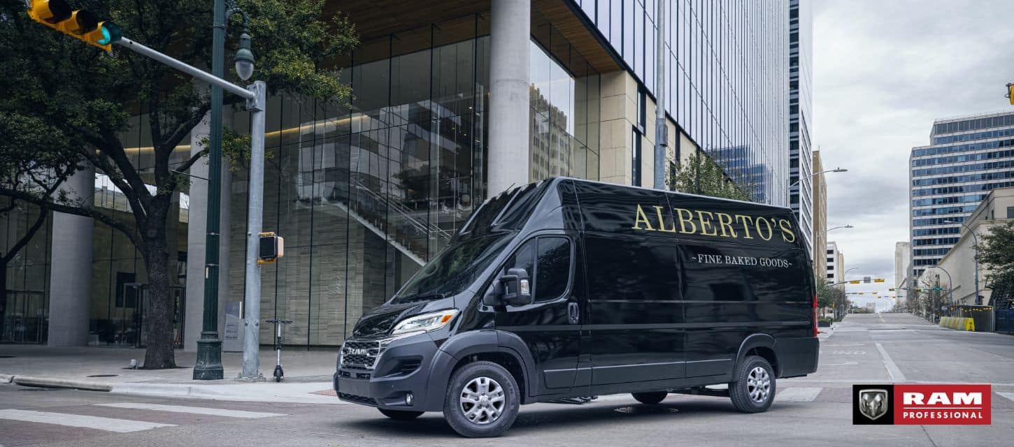 A black 2024 Ram 3500 ProMaster SLT+ Cargo Van Super High Roof with bakery signage on its driver-side rear panel, making a right turn at a lighted intersection on a city street.