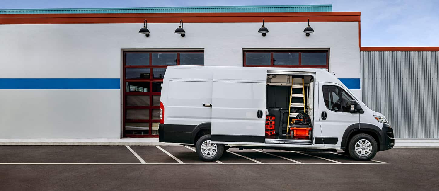 A passenger-side profile of a white Ram ProMaster EV 3500 High Roof Cargo Van parked beside a commercial garage with its sliding side door open, revealing tools, equipment and a ladder inside.