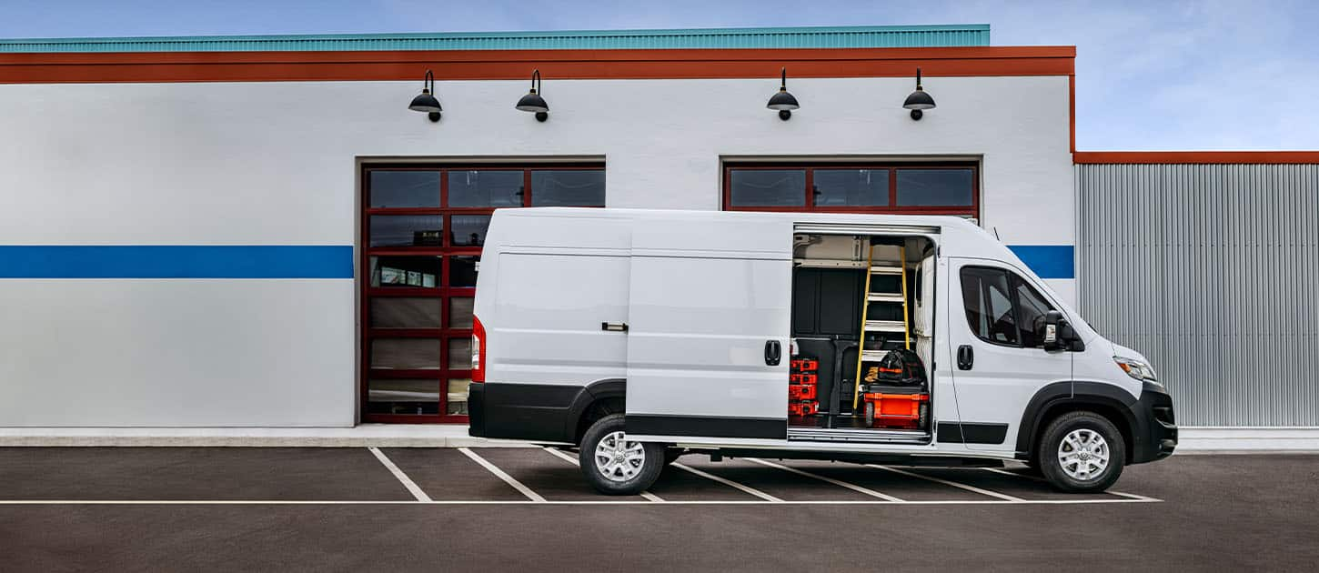 A passenger-side profile of a white Ram ProMaster EV 3500 High Roof Cargo Van parked beside a commercial garage with its sliding side door open, revealing tools, equipment and a ladder inside.