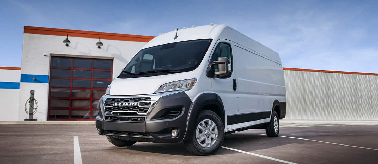An angled driver-side front of a white Ram ProMaster EV 3500 High Roof Cargo Van parked near a commercial garage with a charging station just outside the garage door.