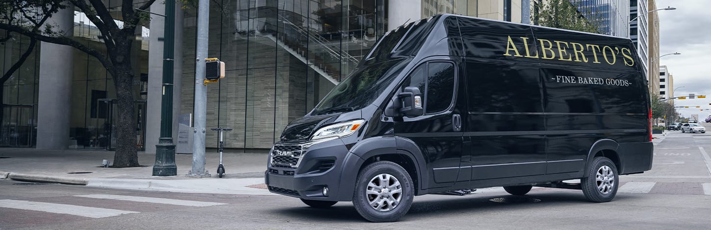 A black 2024 Ram ProMaster SLT+ Cargo Van Super High Roof  with bakery signage on its driver-side rear panel, making a right turn at a lighted intersection on a city street.