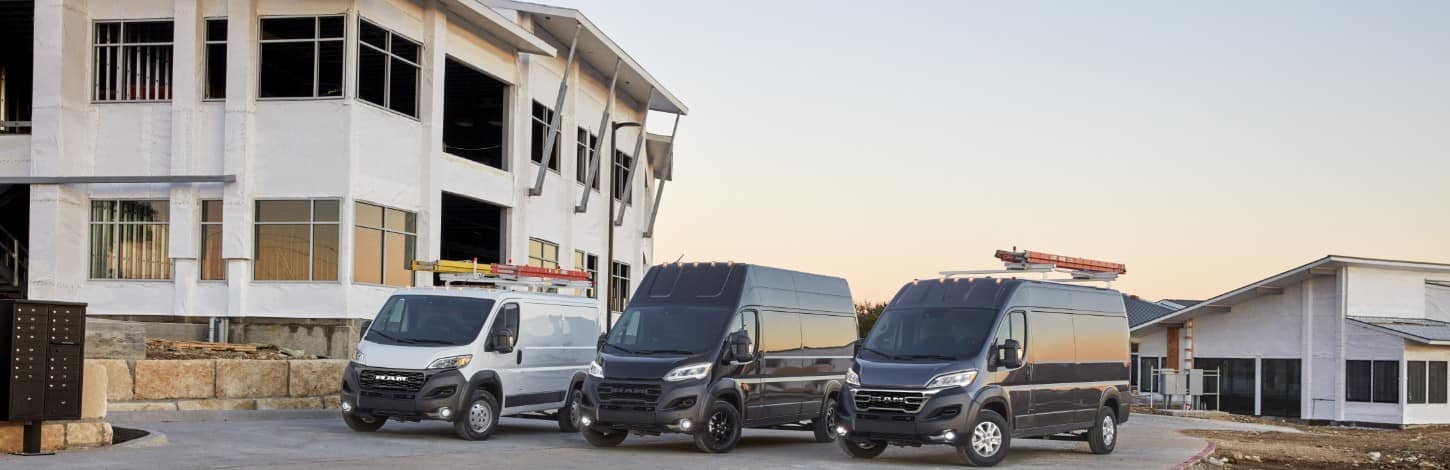 A lineup of three 2024 Ram ProMaster Vans parked at a construction site, from left to right: a white 1500 Tradesman Cargo Van Standard Roof with a ladder on its roof rack, a black 3500 SLT Plus  Cargo Van Super High Roof and a black 2500/3500 SLT Cargo Van High Roof with a ladder on its roof rack.