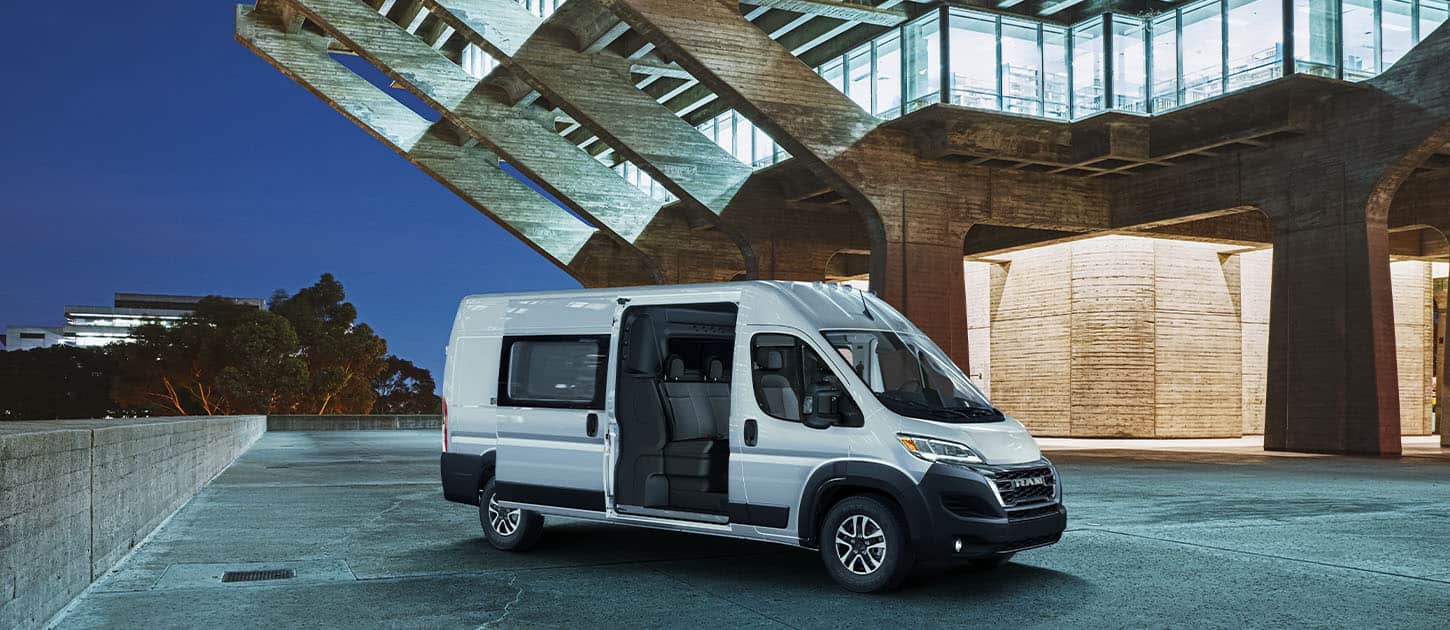 An angled passenger-side profile of a white 2024 Ram ProMaster 2500 SLT Crew Van High Roof parked next to a contemporary building at night, with its passenger-side sliding door open, revealing a second row of seats. 