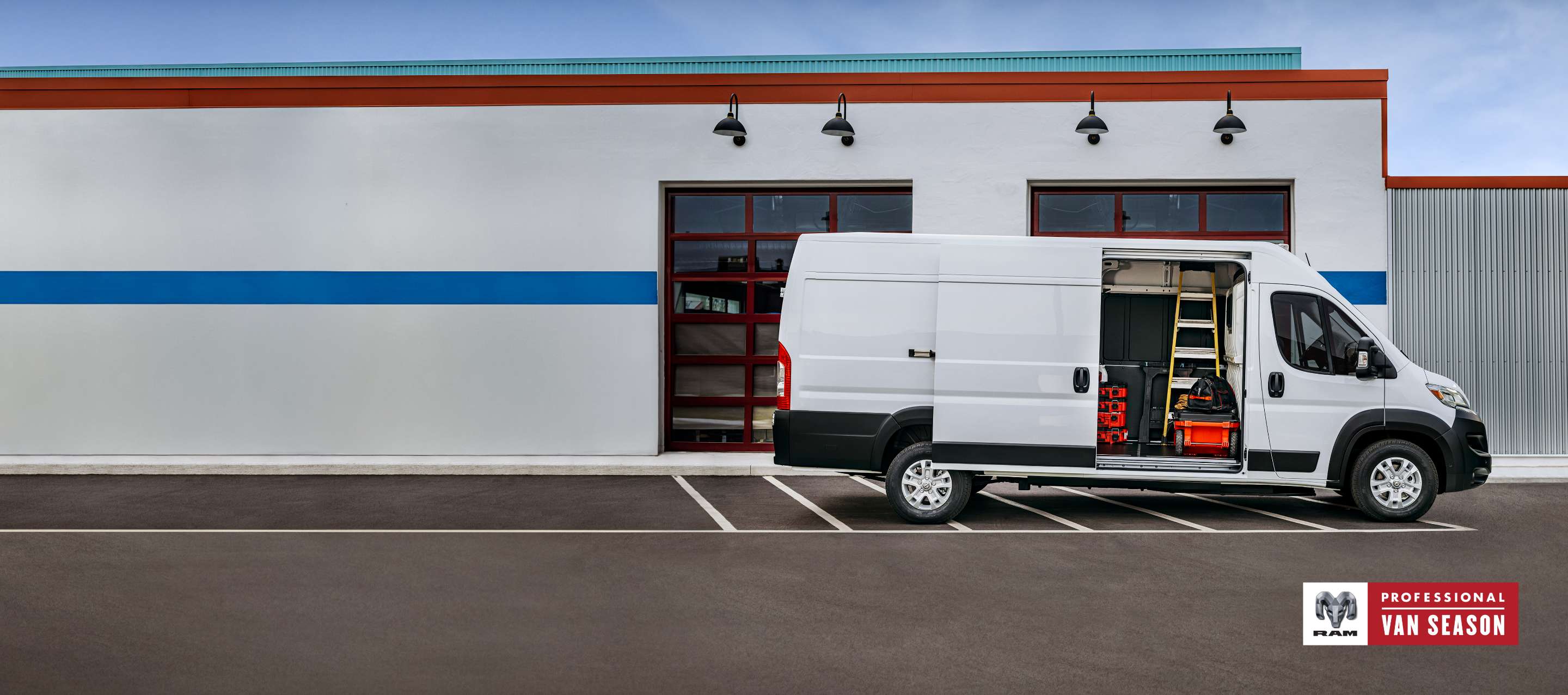 A passenger-side profile of a white 2024 Ram ProMaster EV High Roof Cargo Van (Preproduction Cargo Van shown. Actual production vehicle and feature may vary), parked beside a commercial garage with its passenger-side sliding door open revealing a ladder, tools and equipment inside. Professional Van Season.
