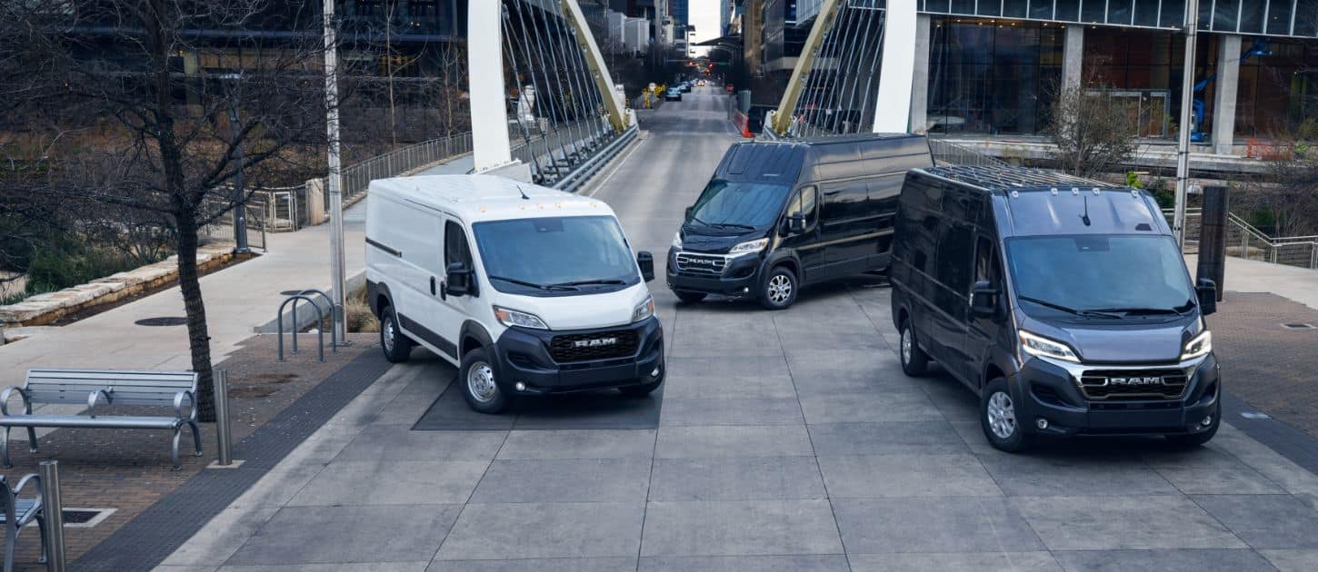 Three 2024 Ram ProMaster models, from left to right: a white Ram ProMaster 1500 Tradesman Cargo Van Standard Roof, a black Ram ProMaster 3500 SLT Plus Cargo Van Super High Roof and a black Ram ProMaster 3500 SLT Cargo Van High Roof parked just beyond a bridge in the center of a busy large city.