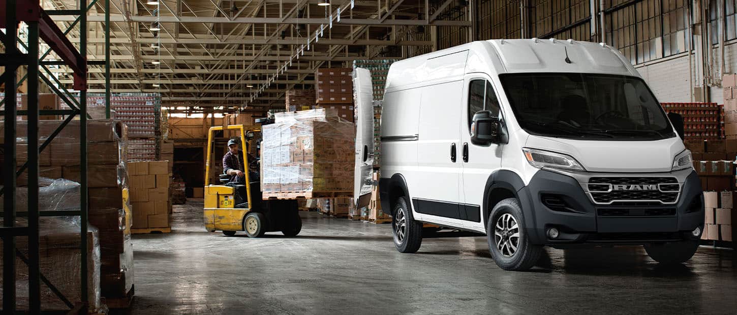 An angled front profile of a white 2024 Ram ProMaster 2500 SLT Cargo Van High Roof, with its rear doors fully open and a forklift with a pile of boxes being driven toward the back of the van.