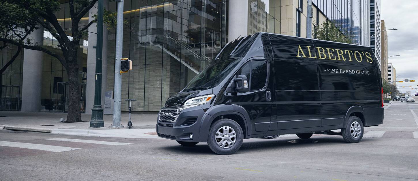 A black 2024 Ram ProMaster 3500 SLT+ Cargo Van Super High Roof with bakery signage on its driver-side rear panel, making a right turn at a lighted intersection on a city street.