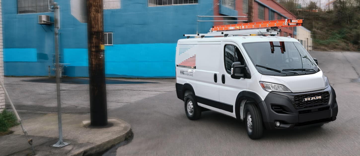 A white 2024 Ram ProMaster Tradesman Cargo Van Standard Roof with the logo for an HVAC company on its side and a ladder on its roof rack making a right turn through an industrial area.