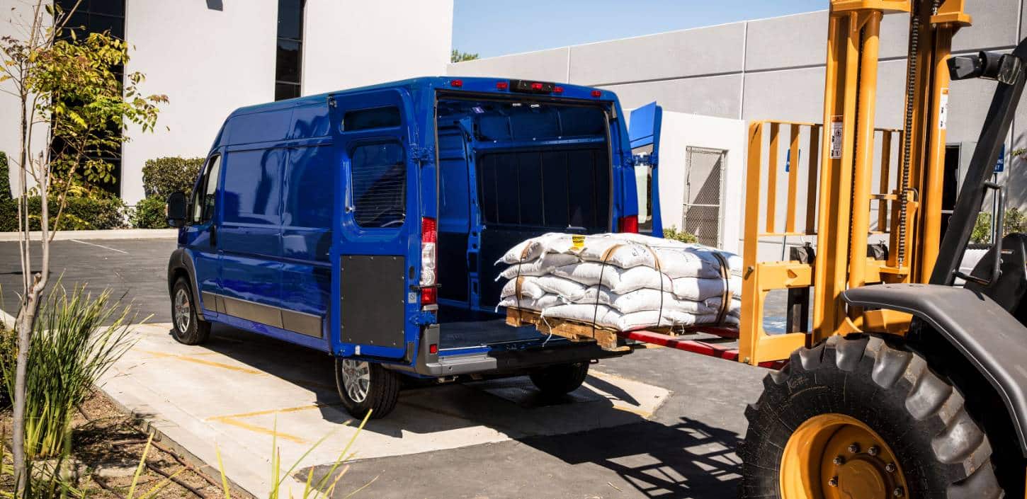Display An angled rear profile of a blue 2024 Ram ProMaster 2500/3500 SLT Cargo Van High Roof, with its rear doors fully open and a pile of bagged materials being loaded into the cargo area by a forklift.