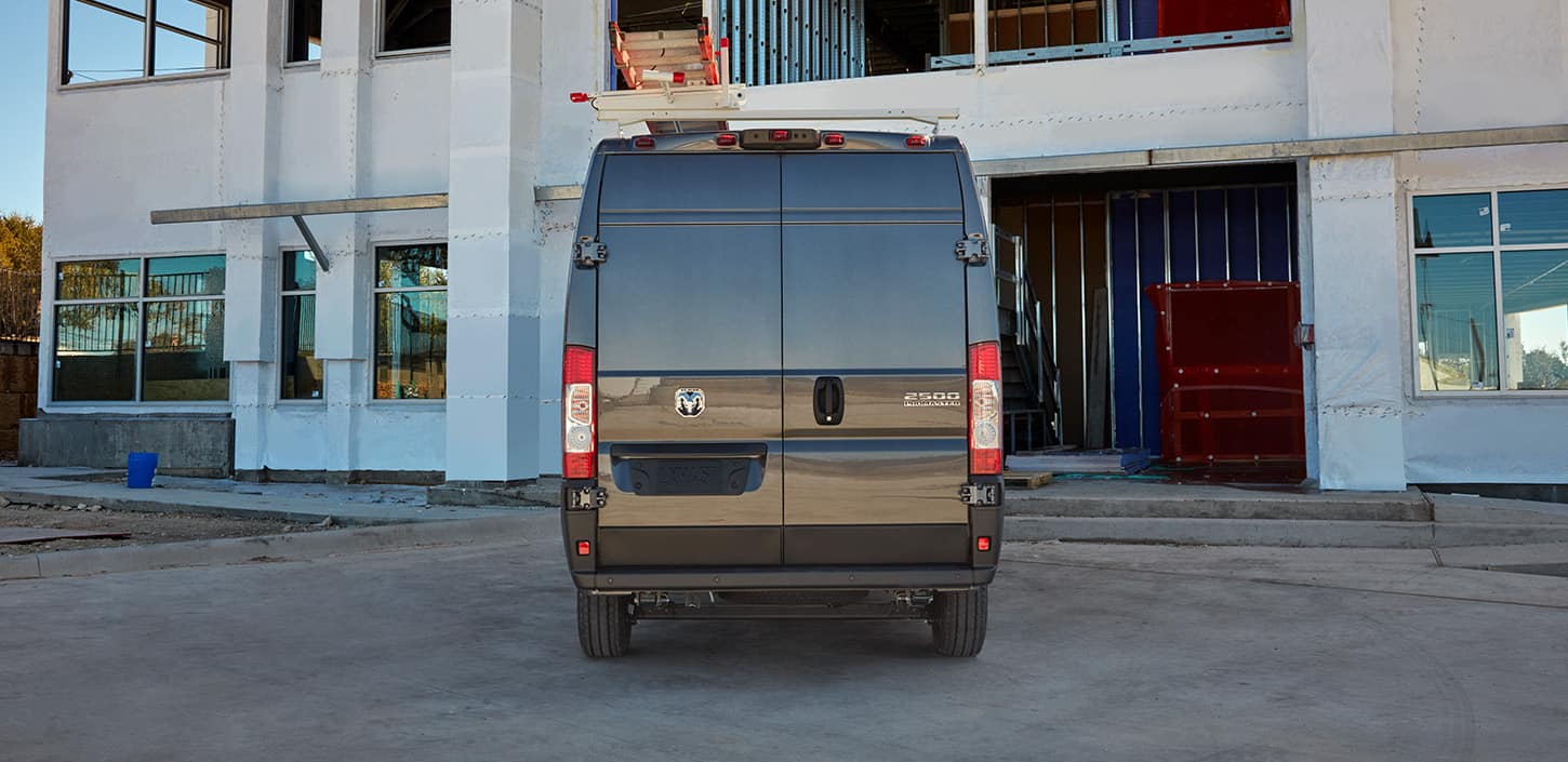 Display A rear profile of a black 2024 Ram ProMaster 2500 Cargo Van High Roof, with a ladder rack, pulling into the garage of a commercial building.