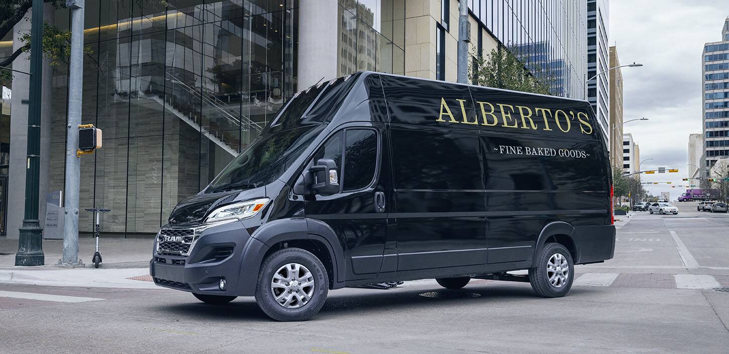 Display A black 2024 Ram ProMaster 3500 SLT+ Cargo Van Super High Roof with bakery signage on its driver-side rear panel, making a right turn at a lighted intersection on a city street.