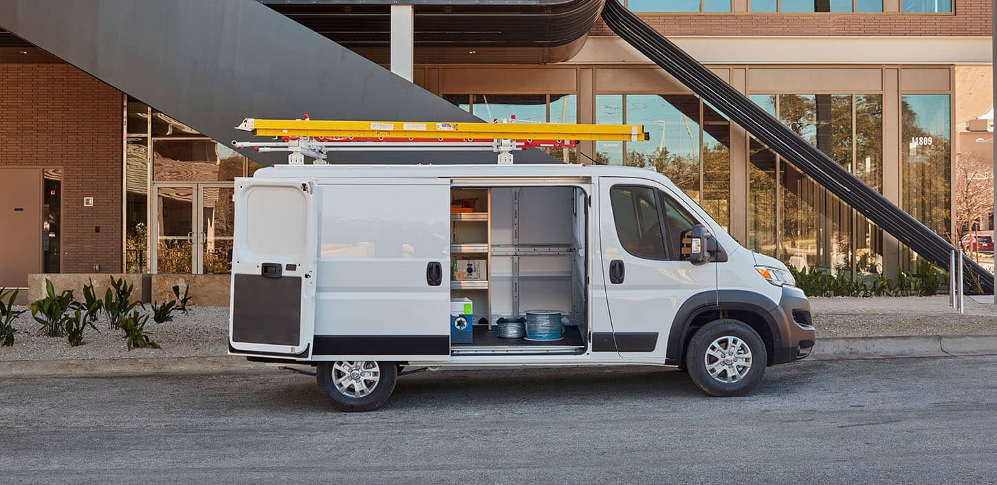A passenger-side profile of a white 2024 Ram ProMaster 2500 SLT Cargo Van High Roof, parked next to a contemporary building with a ladder on its roof rack and its passenger-side sliding door and rear doors open.