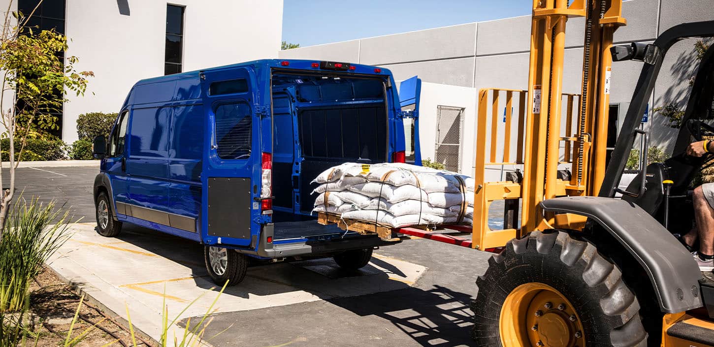 Display An angled rear profile of a blue 2024 Ram ProMaster 2500 SLT Cargo Van High Roof, with its rear doors fully open and a pile of bagged materials being loaded into the cargo area by a forklift.