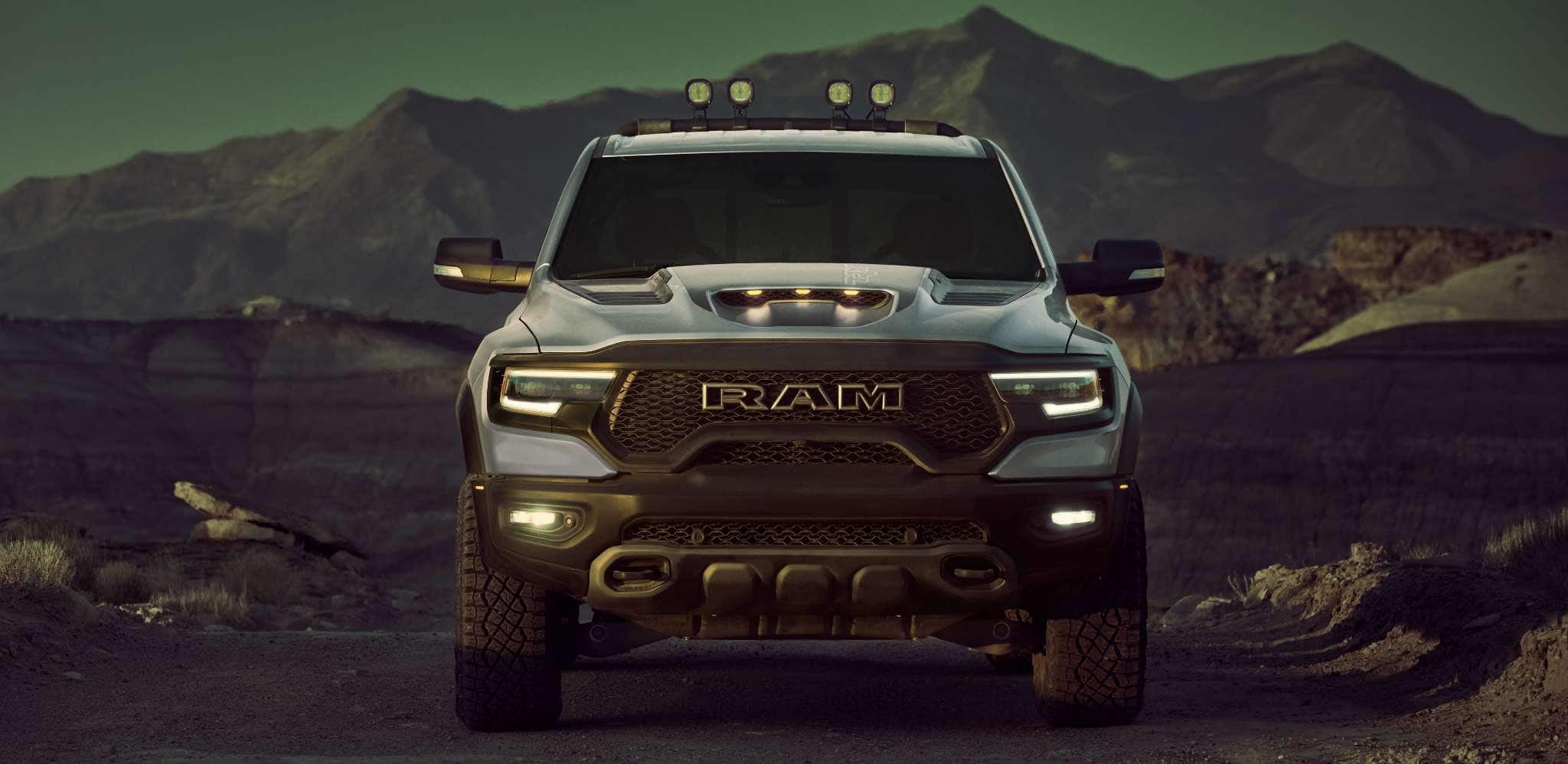 Display A head-on angle of a silver 2024 Ram 1500 TRX Crew Cab with its headlamps on, parked in the desert at dusk, with mountains in the background.