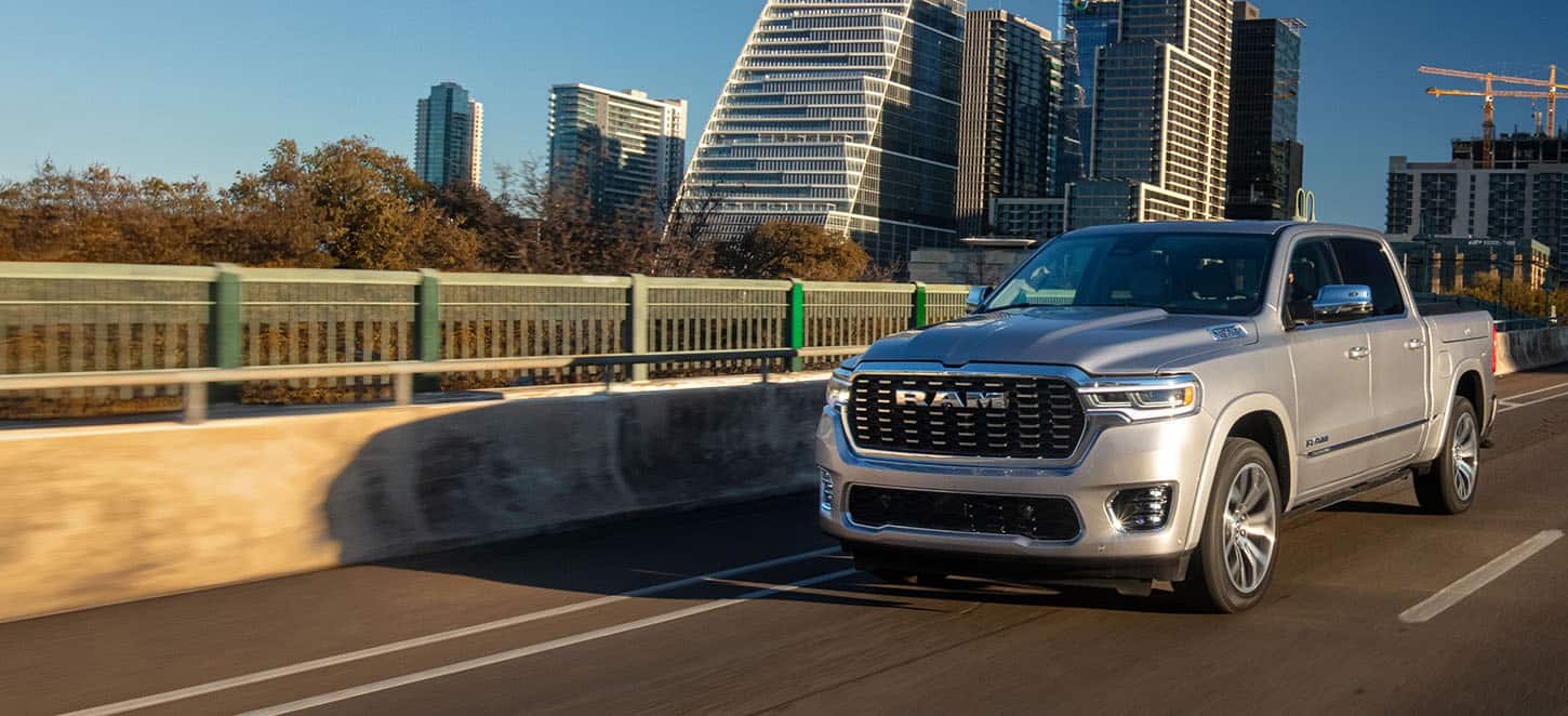 A silver 2025 Ram 1500 Tungsten Crew Cab being driven on a highway bridge in a city, with tall buildings in the background.