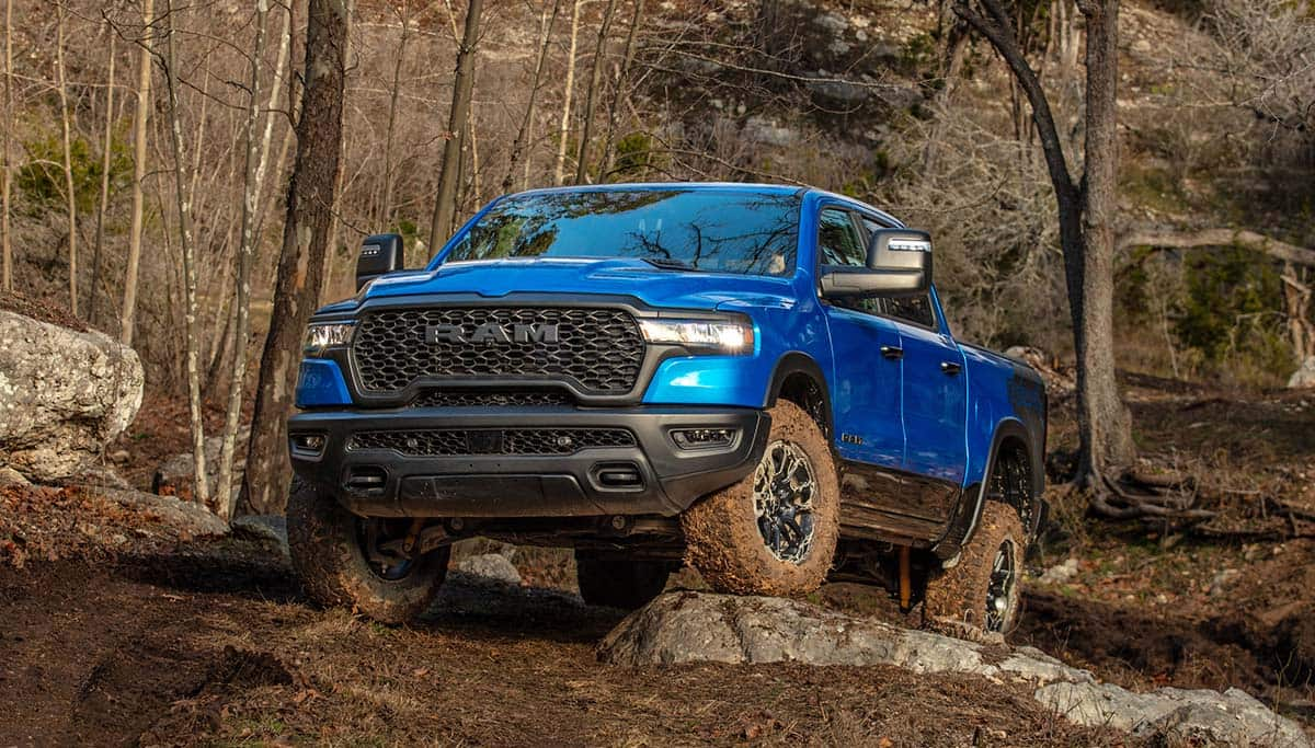 Display A blue 2025 Ram 1500 Rebel Crew Cab climbing a muddy hill off-road.