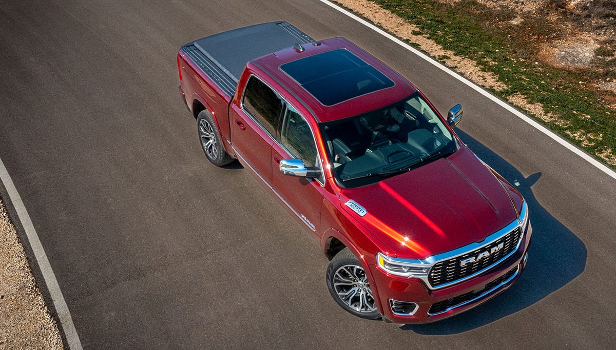 Display A raised angle displaying a red 2025 Ram 1500 Tungsten Crew Cab with sunroof and tonneau cover, being driven on a highway.