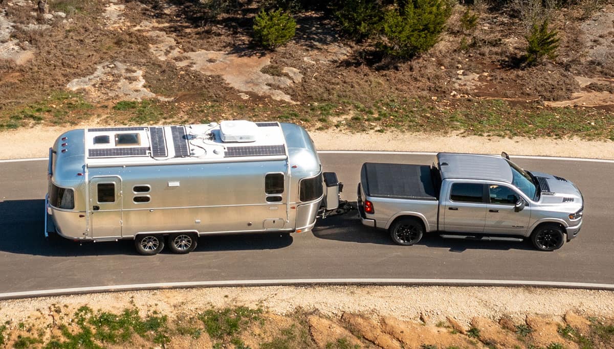 Display A raised angle displaying a silver 2025 Ram 1500 Laramie Crew Cab with tonneau cover, towing a large travel trailer as it's driven down a highway.