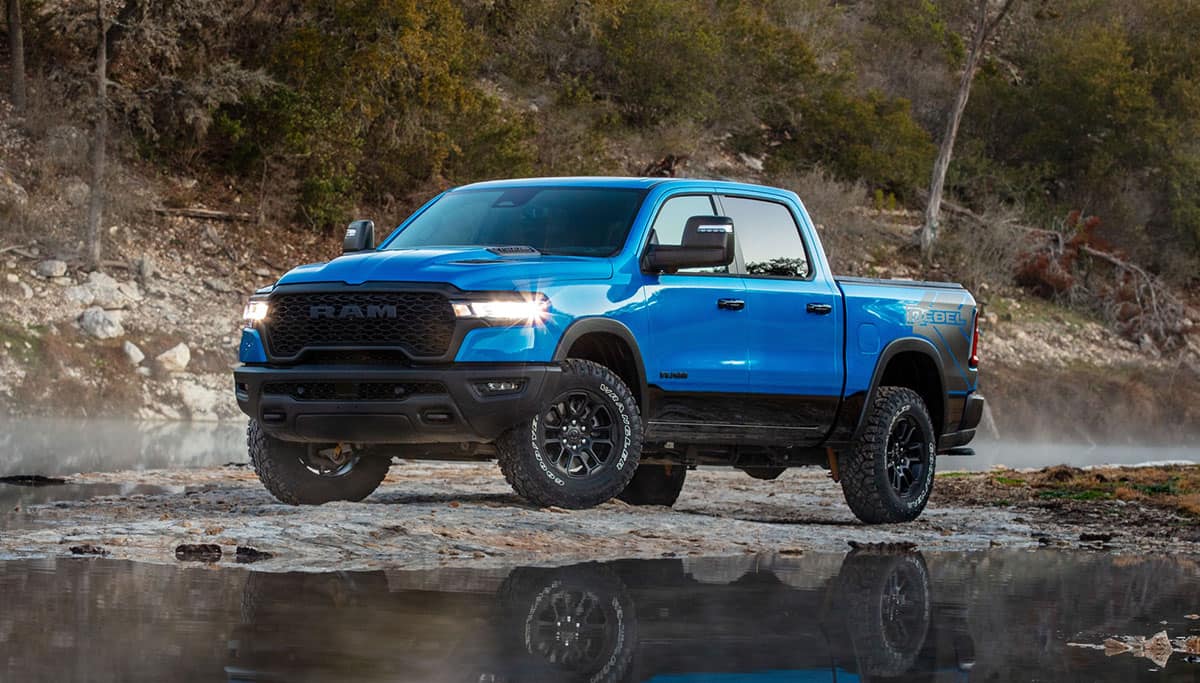 Display A blue 2025 Ram 1500 Rebel Crew Cab being driven on a muddy trail off-road, beside a large hill.