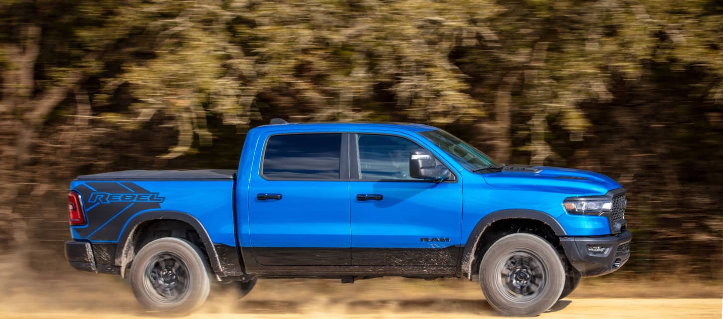 A passenger-side profile of a blue 2025 Ram 1500 Rebel Crew Cab with the background blurred to indicate the vehicle is in motion.